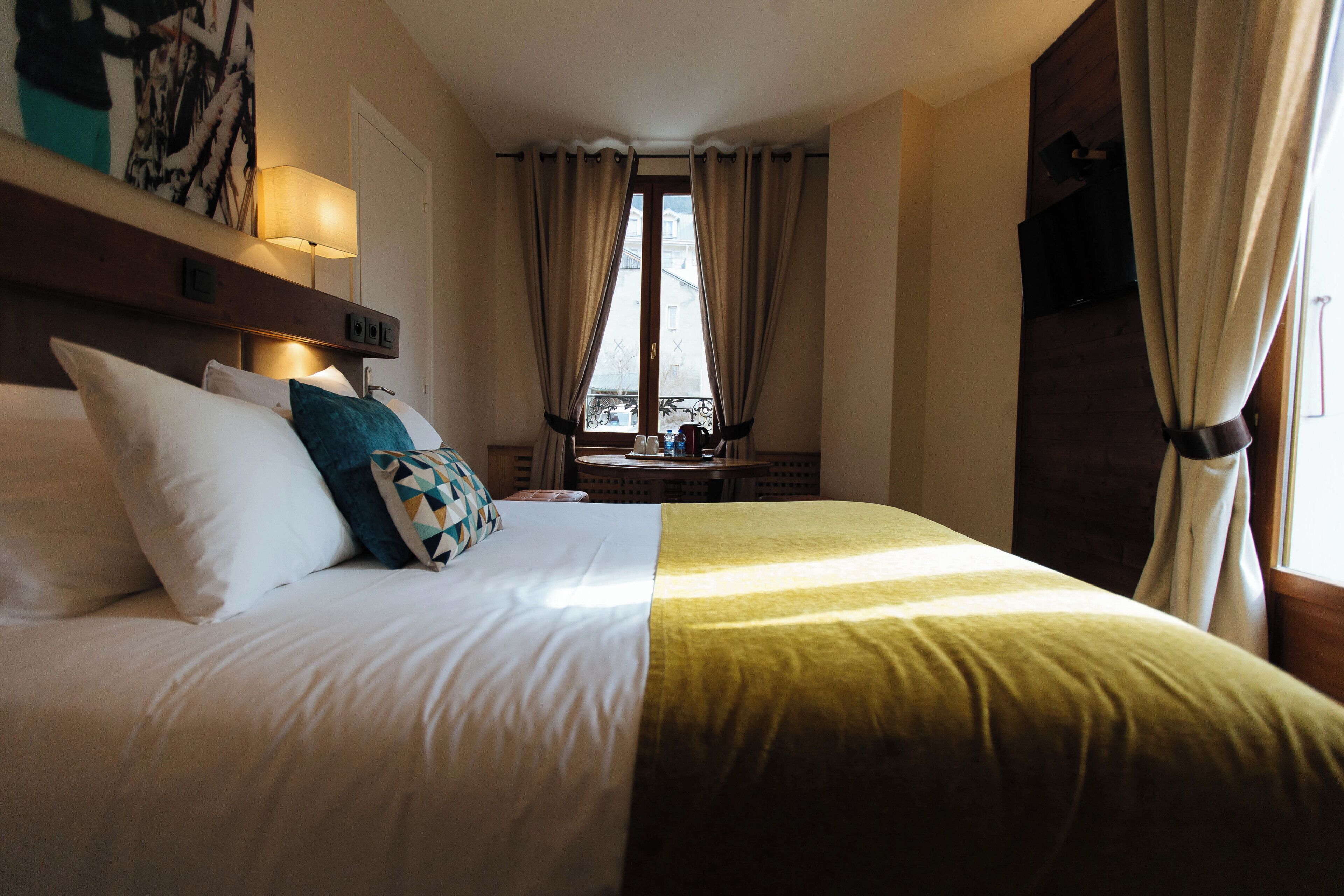 Habitación de hotel con una cama bien hecha, una mesita de noche con lámpara y vistas al exterior a través de amplios ventanales.
