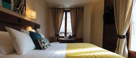 Habitación de hotel con una cama bien hecha, una mesita de noche con lámpara y vistas al exterior a través de amplios ventanales.