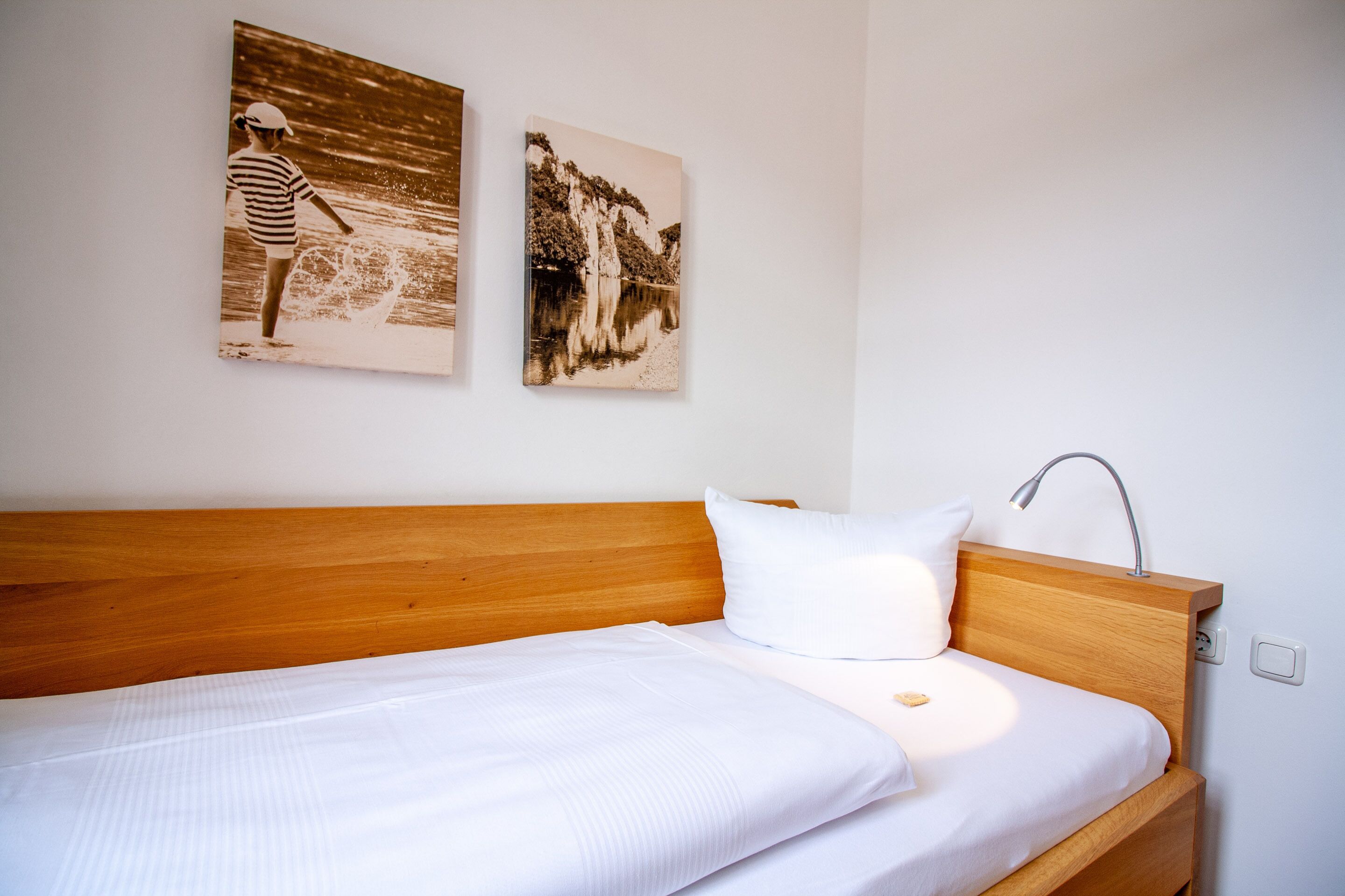 A neatly made bed with a wooden headboard, a white pillow, and a wall-mounted lamp. Two monochrome photographs are hung above the bed.