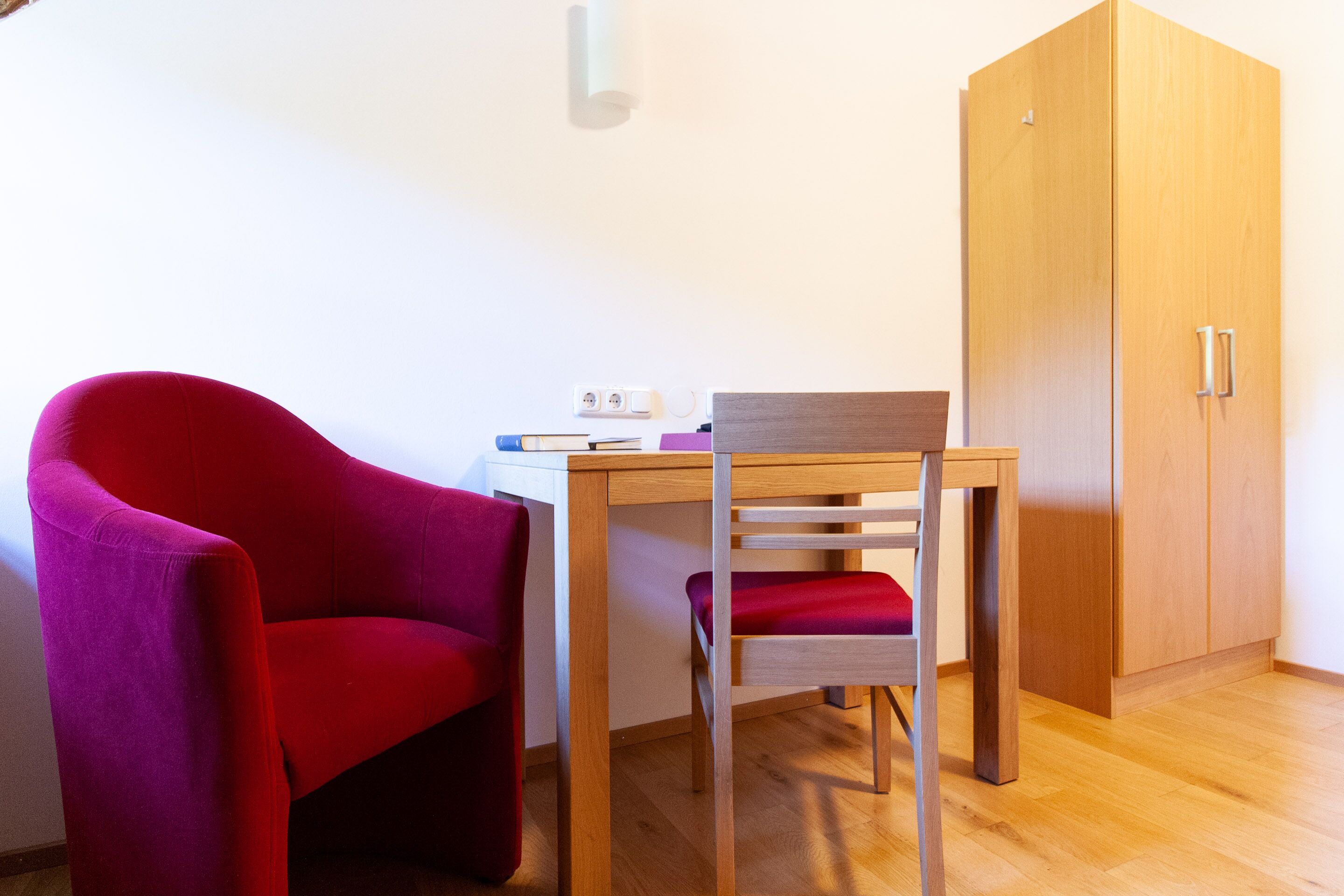 A room with a wooden desk, a red chair, a wardrobe, and a wall socket.
