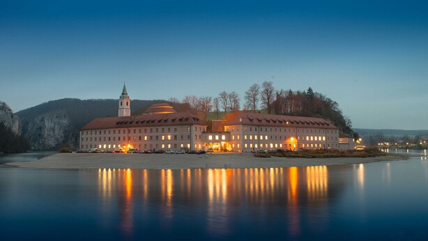 A large, historic building with a prominent tower, situated by a calm body of water, reflecting its lights.
