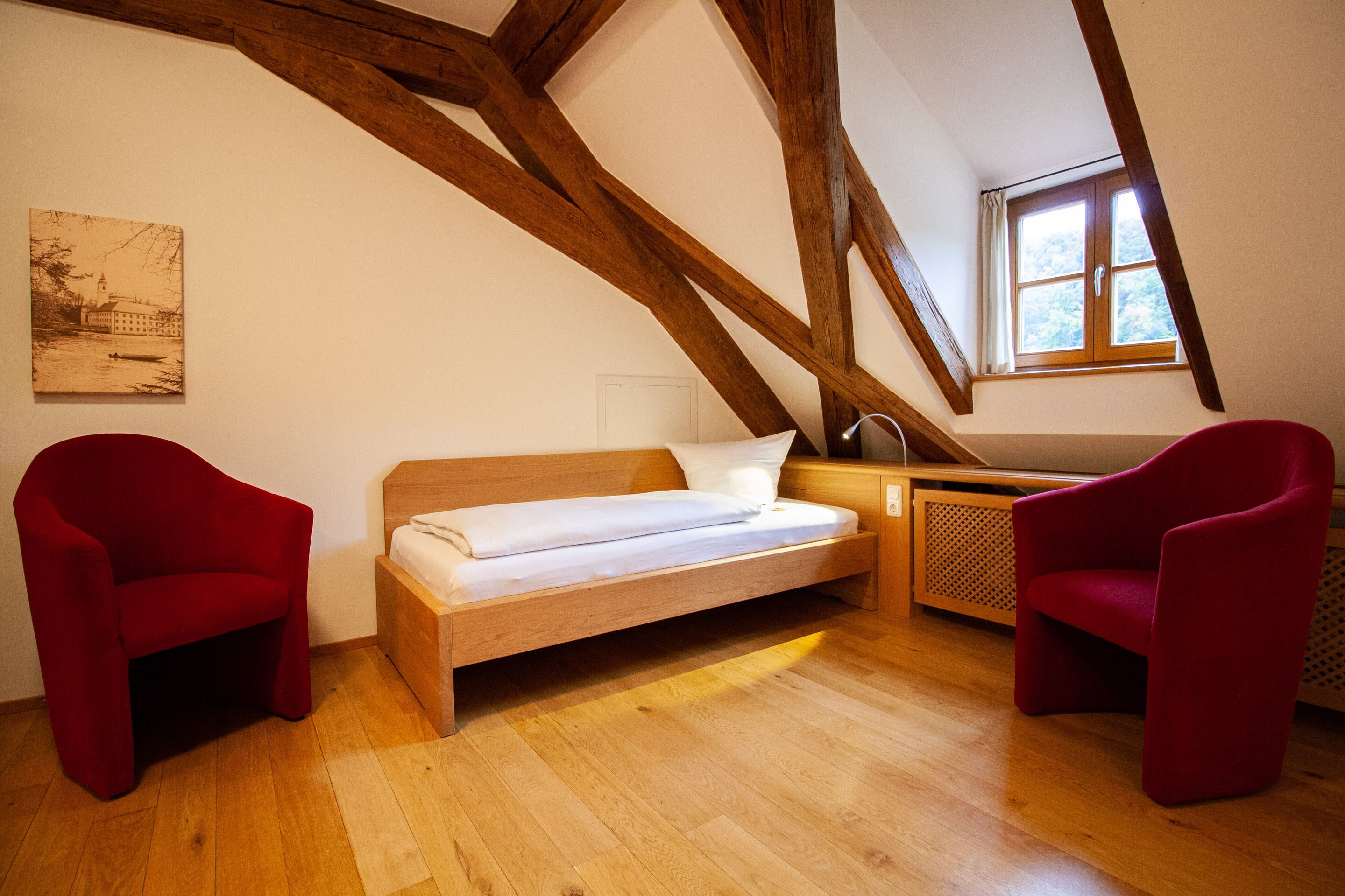 A loft bedroom with a wooden beam ceiling, a bed, two red chairs, a window, and a painting on the wall.