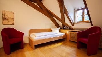 A loft bedroom with a wooden beam ceiling, a bed, two red chairs, a window, and a painting on the wall.