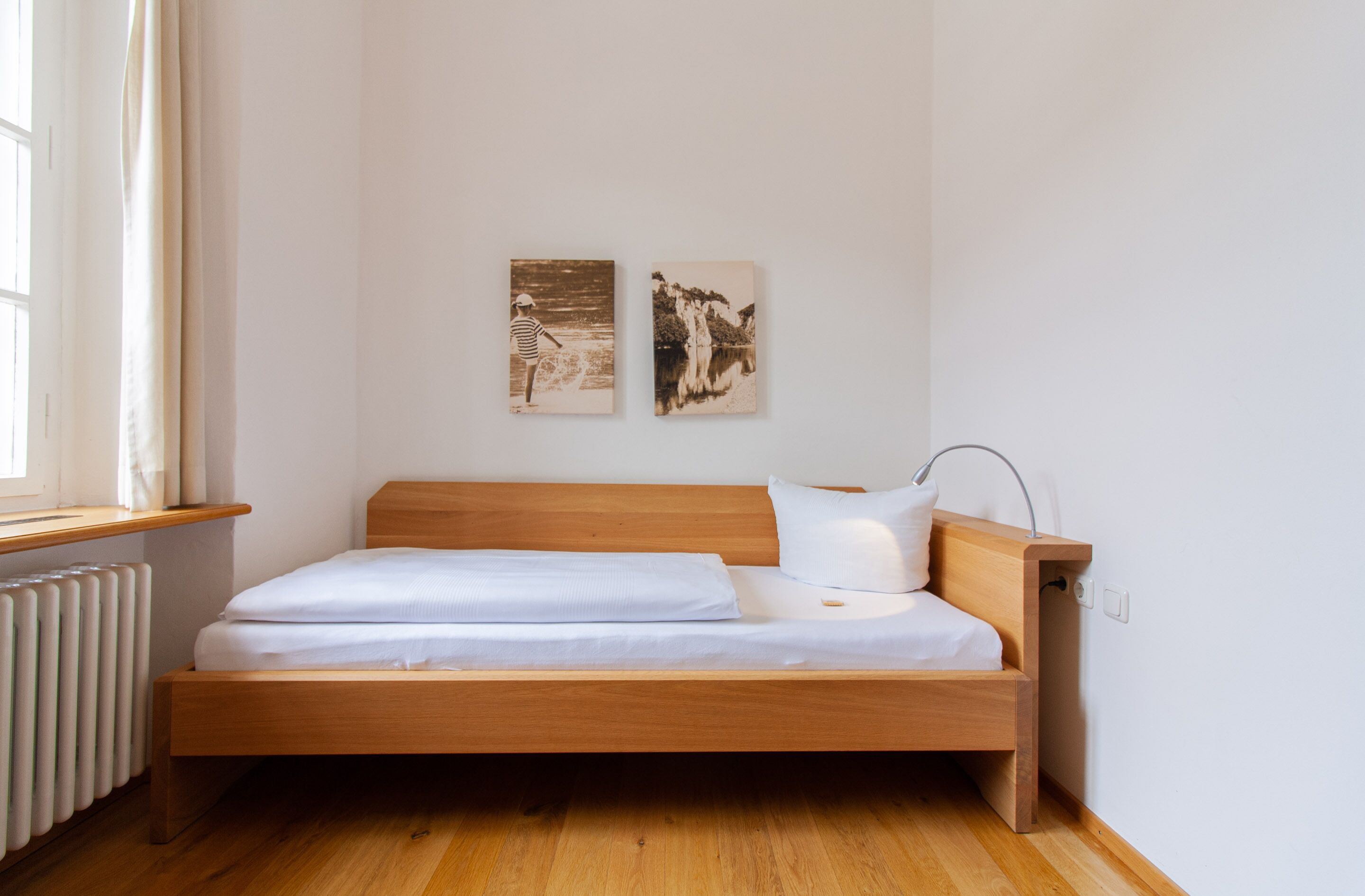 A single bed with a wooden frame, white bedding, and a bedside table with a lamp. There are two framed pictures on the wall above the bed.