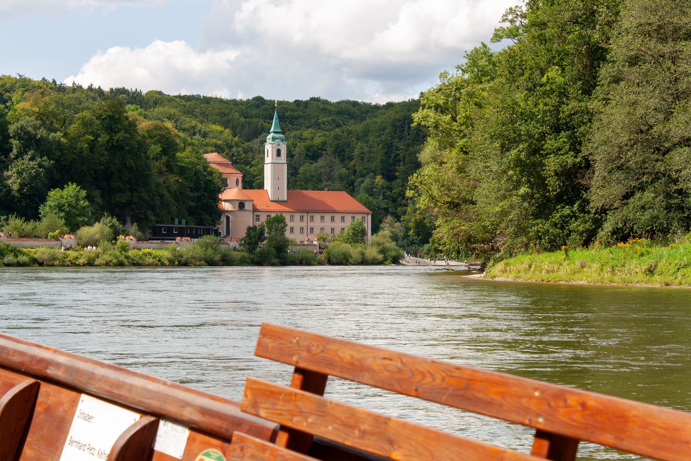 A riverside view of a historic building with a tower, surrounded by lush greenery.