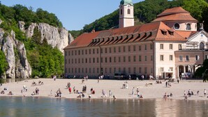 A historic building with a clock tower, situated by a river with a rocky cliff in the background.