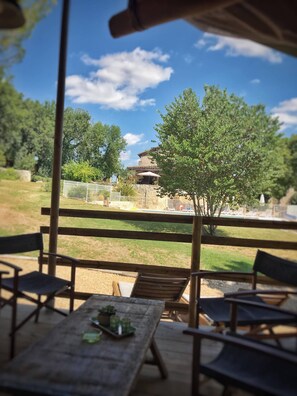 Een terras met houten tafels en stoelen, uitzicht op een gebouw met pannendak, en een omheind gebied met bomen en een zwembad op de achtergrond.