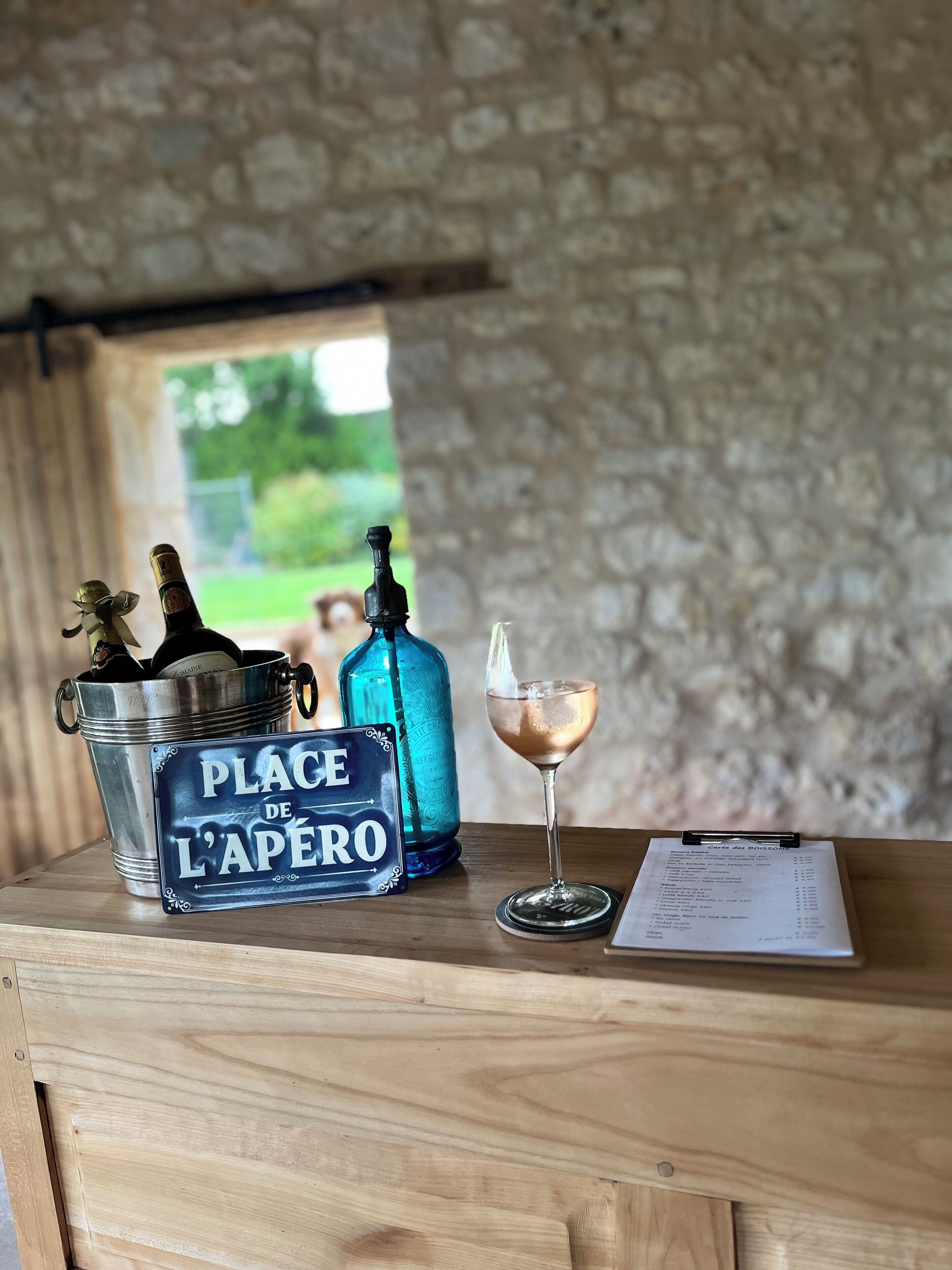A wooden table with a metal bucket labeled "PLACE DE L'APÉRO," a blue bottle, a wine glass with a drink, and a menu on a coaster.