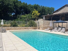 Una piscina con agua azul cristalina, rodeada por un muro de piedra y un solárium con baldosas. Hay tumbonas y sombrillas a lo largo de la piscina.