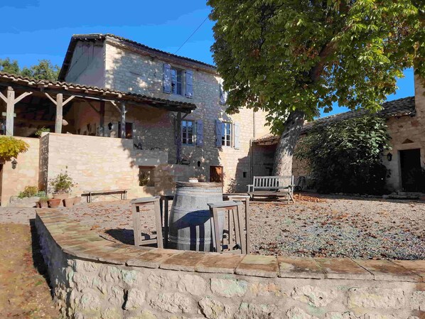 Relax in a tranquil courtyard with a wooden bench, table, and barrel, surrounded by a traditional stone building with a tiled roof and balcony.