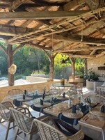 Un espacio de comedor al aire libre con vigas de madera rústicas, mobiliario de mimbre y un fondo de pared de piedra.