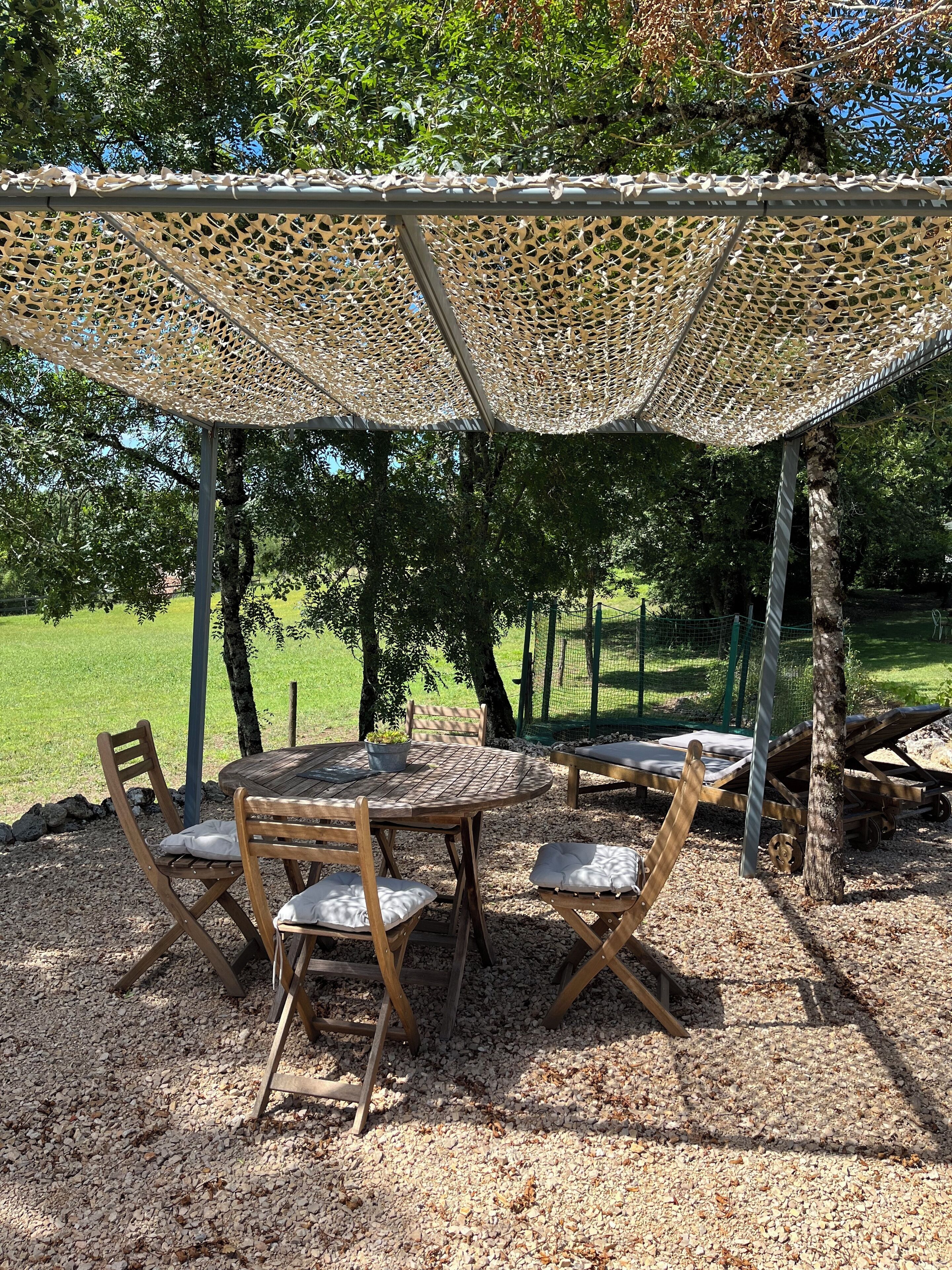 An outdoor seating area with a round table and chairs, covered by a woven shade, surrounded by trees and gravel.