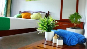 Una cama bien tendida con ropa de cama verde y blanca, una planta en maceta, una toalla azul y una botella pequeña sobre una mesa de madera en una habitación con estructura de cama de madera y un espejo.
