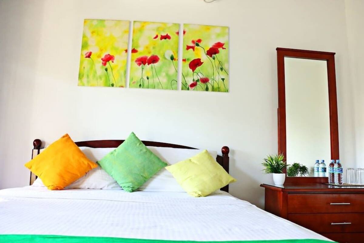 Habitación de hotel con una cama, tres almohadas, una mesita de noche con un espejo y un cuadro colgado en la pared de flores rojas.