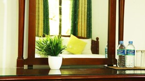 Un aparador de madera con un espejo que refleja una cama con un cojín amarillo, una planta verde en una maceta blanca y dos botellas de agua.