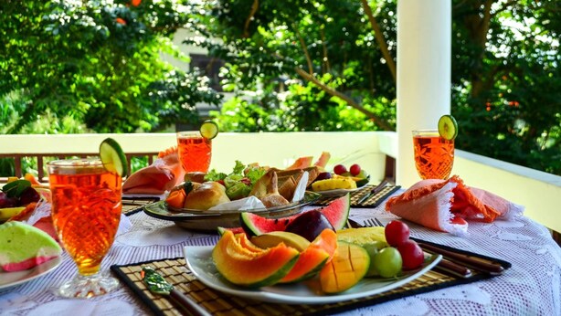 Una mesa con varias frutas, un vaso de té helado y un plato de comida en un balcón con un fondo verde exuberante.