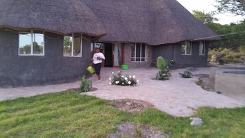 Una casa con techo de paja, un acceso empedrado, un pequeño jardín con flores blancas y una planta de cactus.