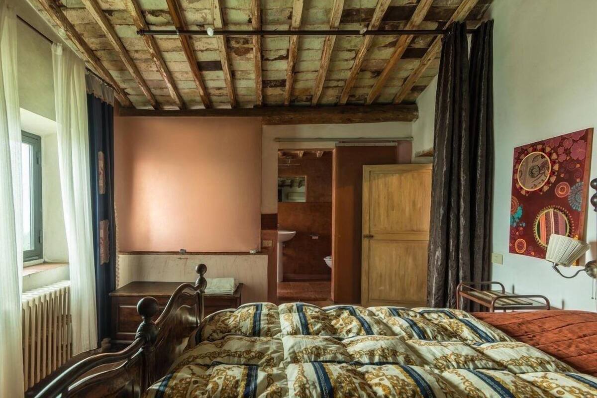 A bedroom with a wooden ceiling, a bed with patterned bedding, a wooden desk, a radiator, and a window with curtains.