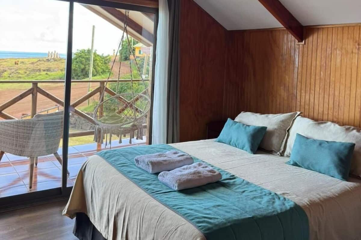 A bedroom with a large bed, a view of the outdoors, and a wooden ceiling.