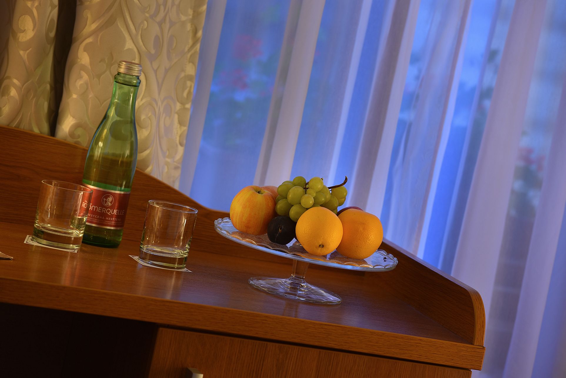 Una mesa de madera con una botella de agua, dos copas para shots y un plato de frutas con naranjas, uvas y una ciruela.