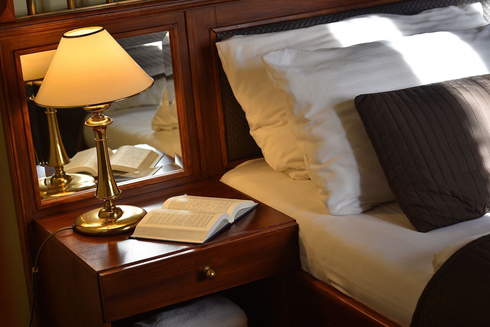 Una cama bien hecha con almohadas blancas y oscuras, una lámpara de noche y un libro abierto sobre una mesita de madera.