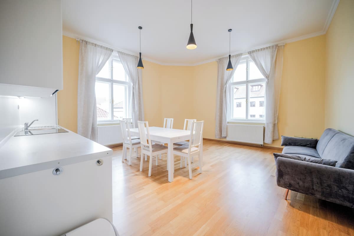 Una cocina moderna con encimera blanca y un lavamanos, un comedor con mesa y sillas blancas, y un sofá gris en una habitación con suelo de madera y ventanas amplias.