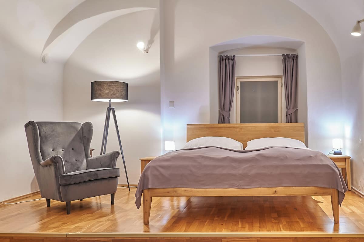 Un dormitorio con una cama de madera, un sillón gris, una lámpara de pie y una mesita de noche.