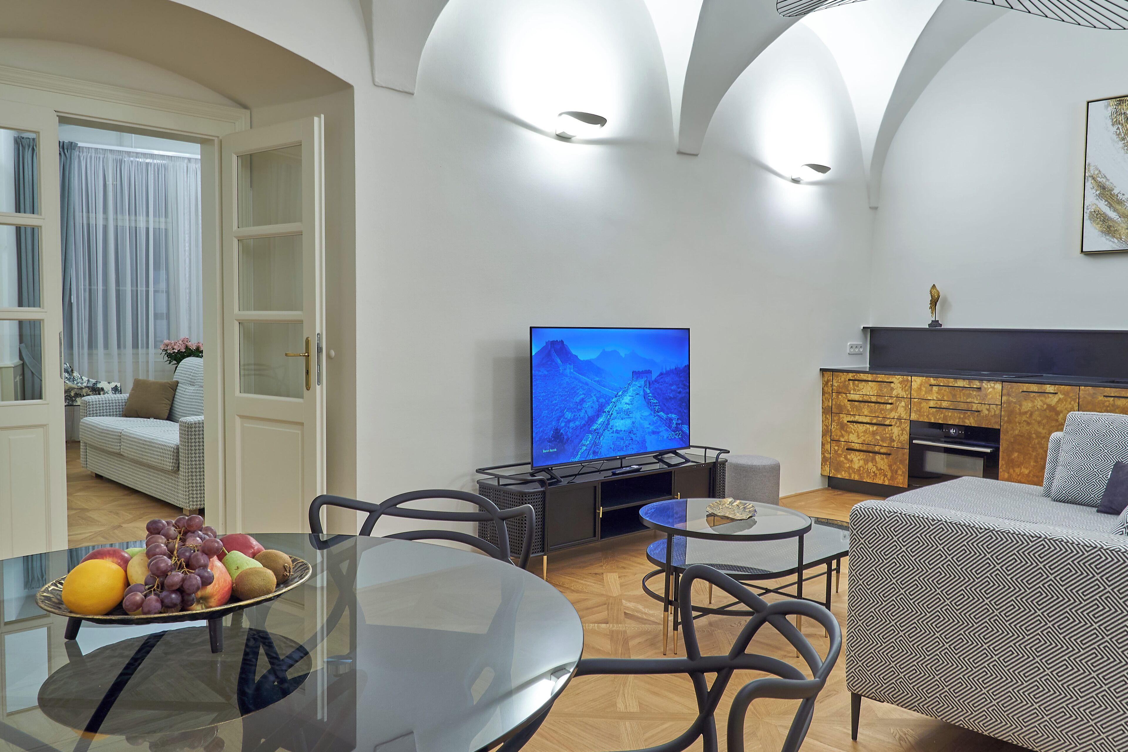 A modern living room with a glass table, a sofa, a television, and a kitchen area with wooden cabinets.