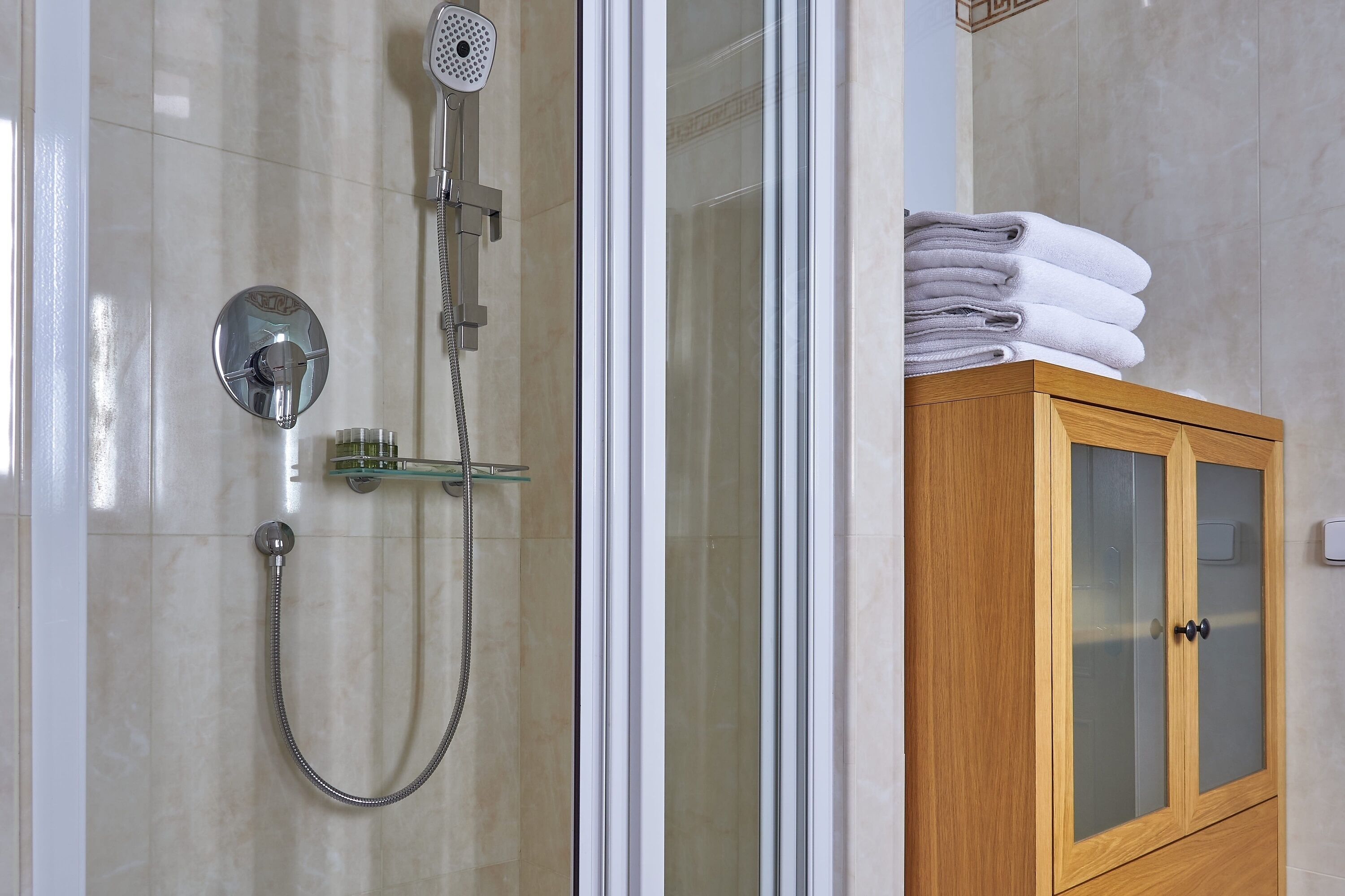 Une douche équipée d’un pommeau de douche à main et d’un robinet mural, un meuble en bois avec des portes en verre, et une pile de serviettes pliées.