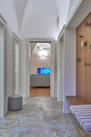 Um corredor com piso estampado, um banquinho cinza, uma porta de madeira e uma televisão ao fundo.