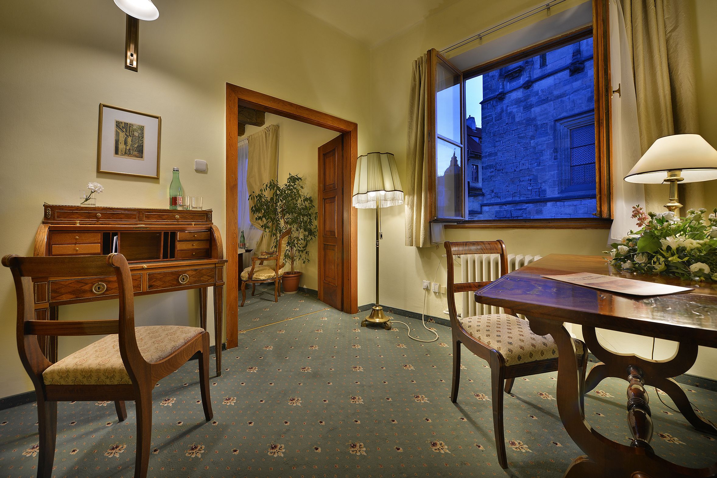 A room with a wooden desk, two chairs, a radiator, a lamp, a framed picture on the wall, and a window with a view of a building.