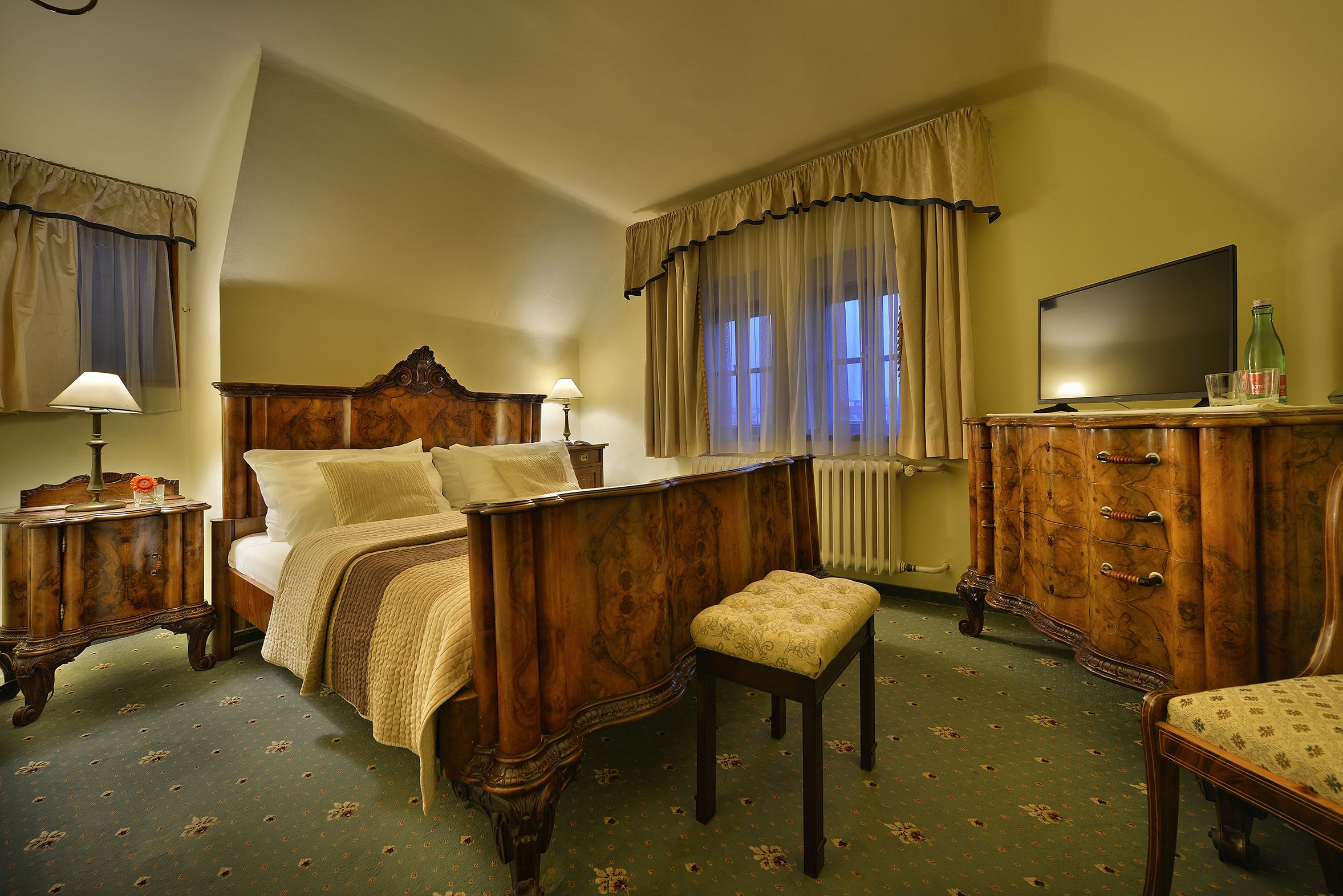 Une chambre à coucher avec un lit en bois, une table de chevet, une commode, un téléviseur et une fenêtre avec des rideaux.