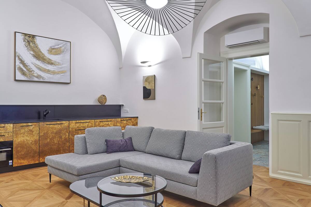 Una sala moderna con un sofá seccional, una mesa de centro de vidrio y una cocina con encimera oscura y armarios de madera.
