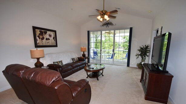 A living room with a brown leather sofa, a coffee table, a ceiling fan, a TV on a wooden stand, and a large window with a view of a balcony. - Ironwood Apartment 1631 (North Myrtle Beach)
