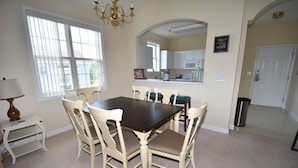 A dining room with a dark wooden table and four chairs, a chandelier, a window with blinds, and a kitchen area in the background.
