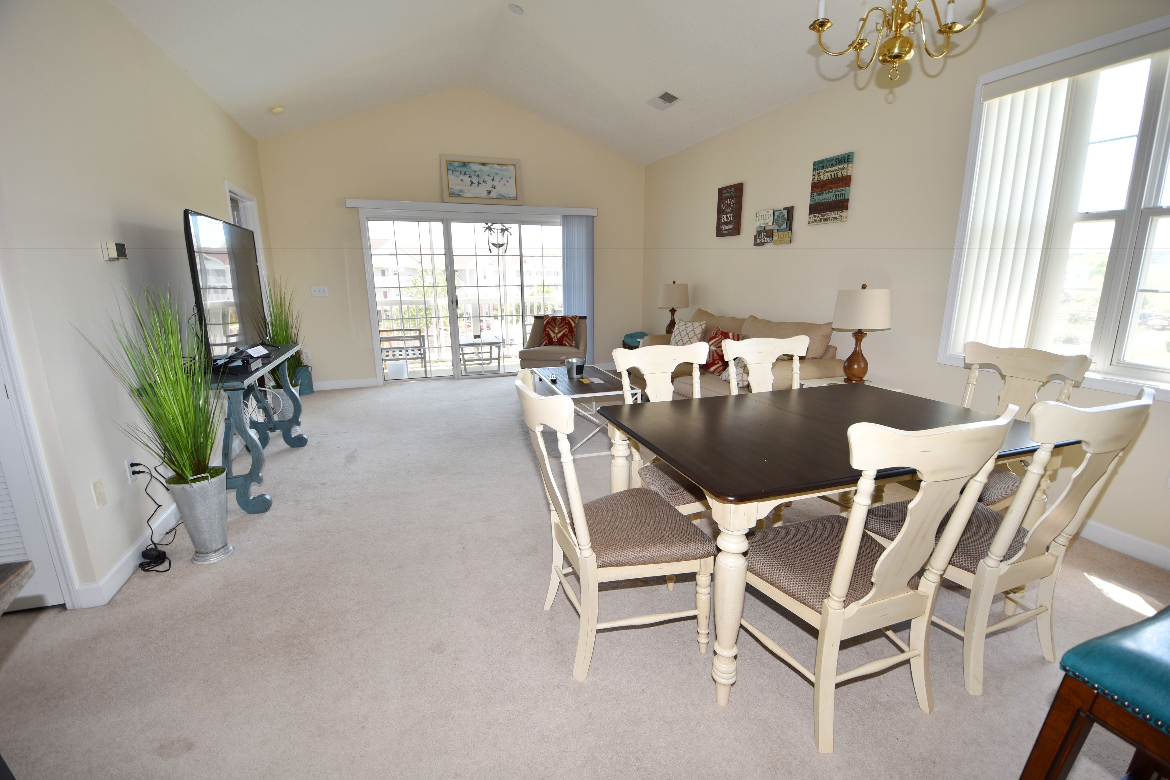 A spacious dining area with a large table and six chairs, a chandelier, and a view through a glass door to the outside.