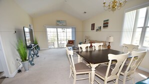 A spacious dining area with a large table and six chairs, a chandelier, and a view through a glass door to the outside.