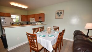 A dining area with a table set for four, surrounded by wooden chairs. A kitchen is visible in the background with wooden cabinets and a refrigerator.