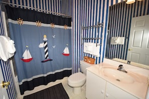 A bathroom with a striped wall, a nautical-themed shower curtain, a white toilet, a sink with a mirror, and a towel rack. - River Crossing Apartment 624 (North Myrtle Beach)