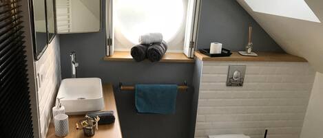 Baño pequeño con lavamanos blanco, inodoro, toallero y ventana con toallas.