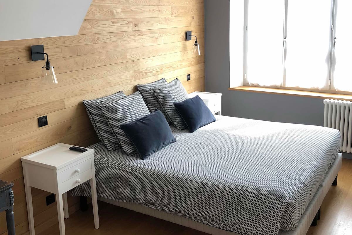 Een slaapkamer met een schuine kap, een bed met een beddegoed met patroon, twee kussens, een nachtkastje met een afstandsbediening en een raam met jaloezieën.