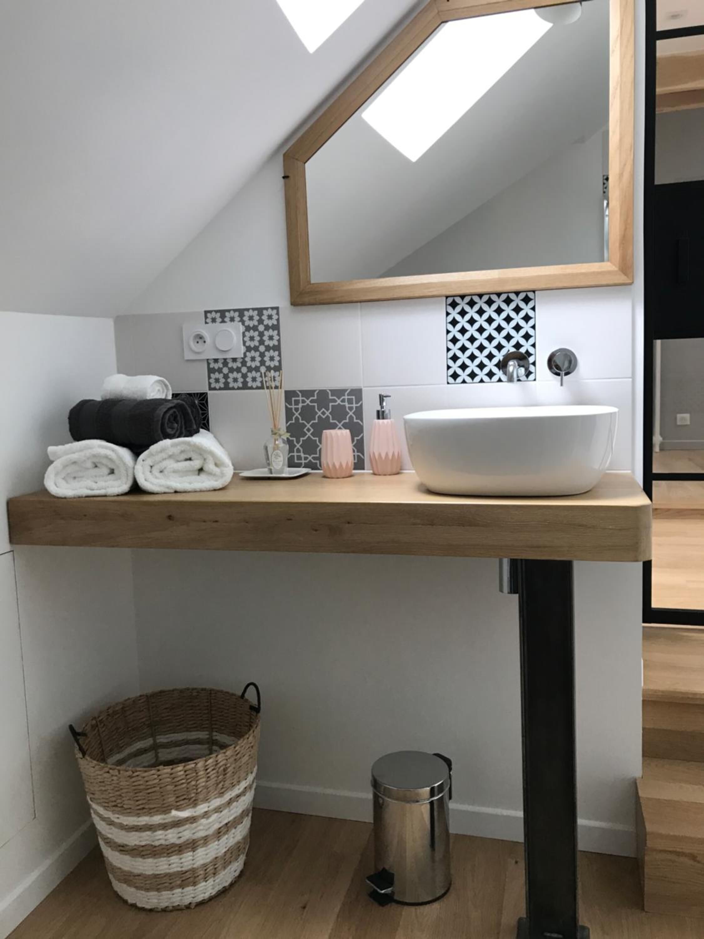 A modern bathroom with a wooden vanity, white sink, and a mirror above. There are towels, a soap dispenser, and a trash can visible.