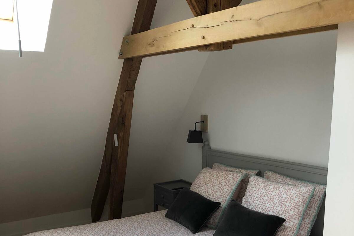 Een slaapkamer met een schuine plafond, een bed met beddengoed met een patroon, een nachtkastje en een wandlamp.