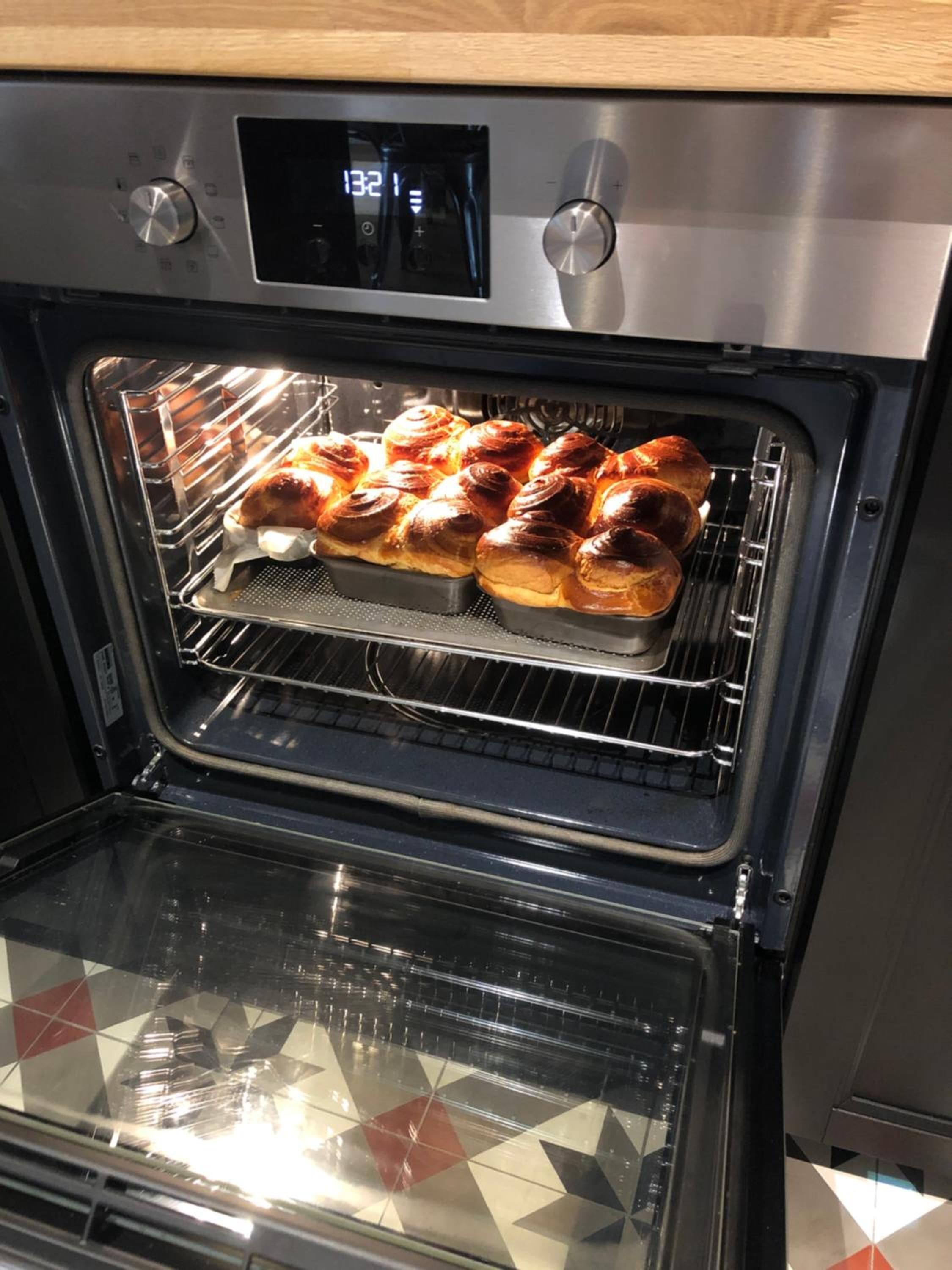 Um forno com visor digital mostrando a hora 13:21. No interior, duas assadeiras com produtos de panificação, possivelmente pão ou doces, no nível do meio.