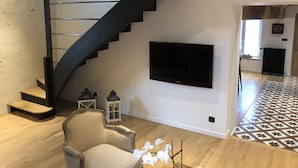 Un salon moderne avec un escalier en colimaçon noir, un téléviseur à écran plat, un fauteuil beige et un sol à motifs.