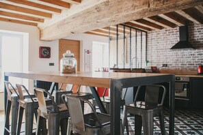 A kitchen with exposed wooden beams, a brick wall, and a large island with chairs. - Chambres d'hôtes Saint Jean (Nogent-le-Rotrou)