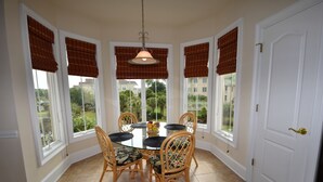 A dining area with a round table and six chairs, large windows with brown blinds, and a white door. - Edgewater Apartment 437 (North Myrtle Beach)