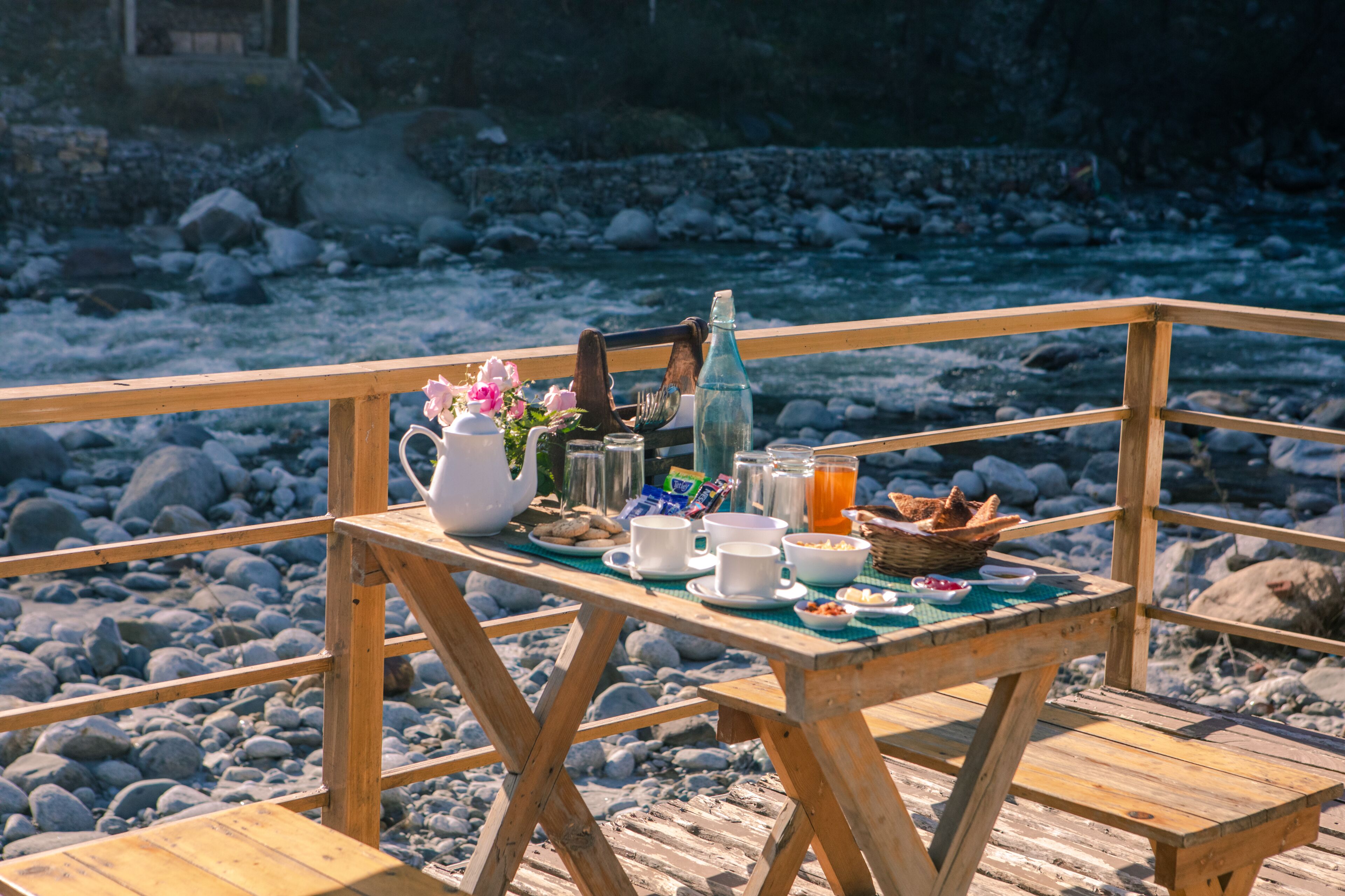 A wooden table set with breakfast items, including bread, eggs, and beverages, on a rocky riverside deck.