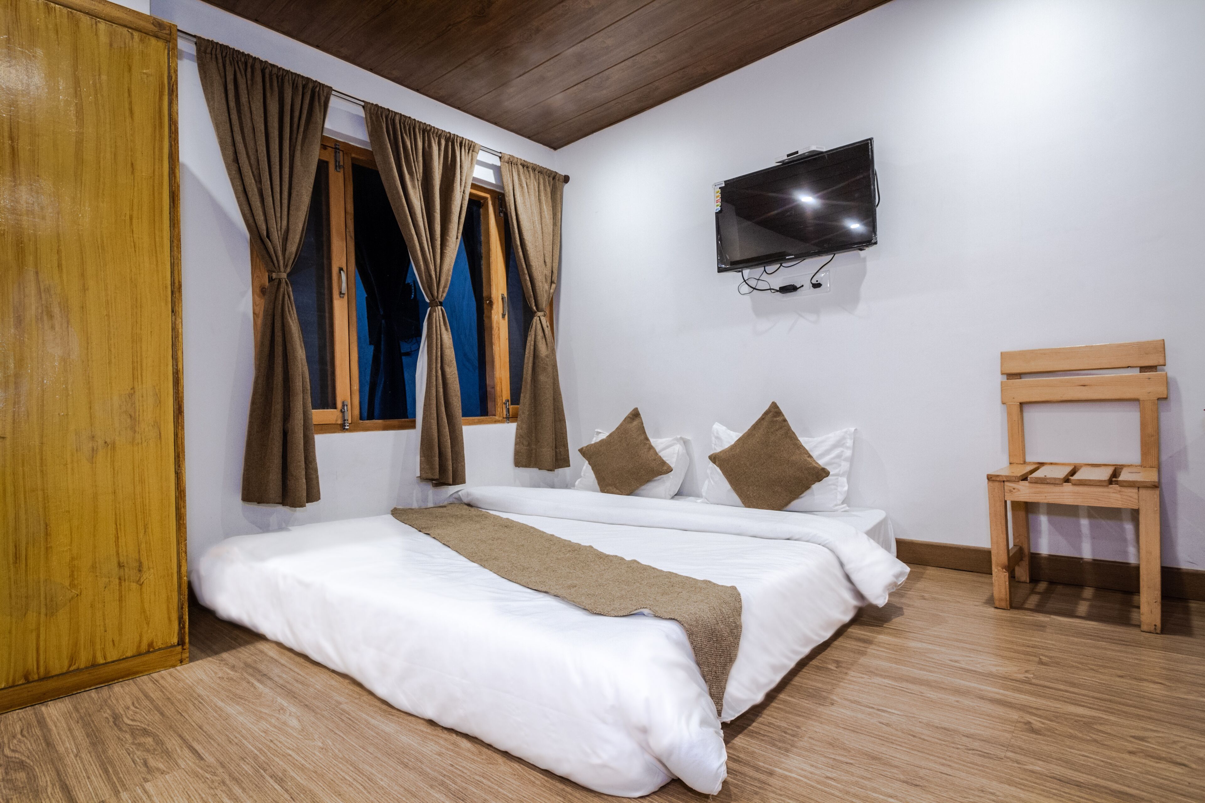 A bedroom with a wooden floor, a bed with white bedding, brown curtains, a wooden chair, and a mounted TV.