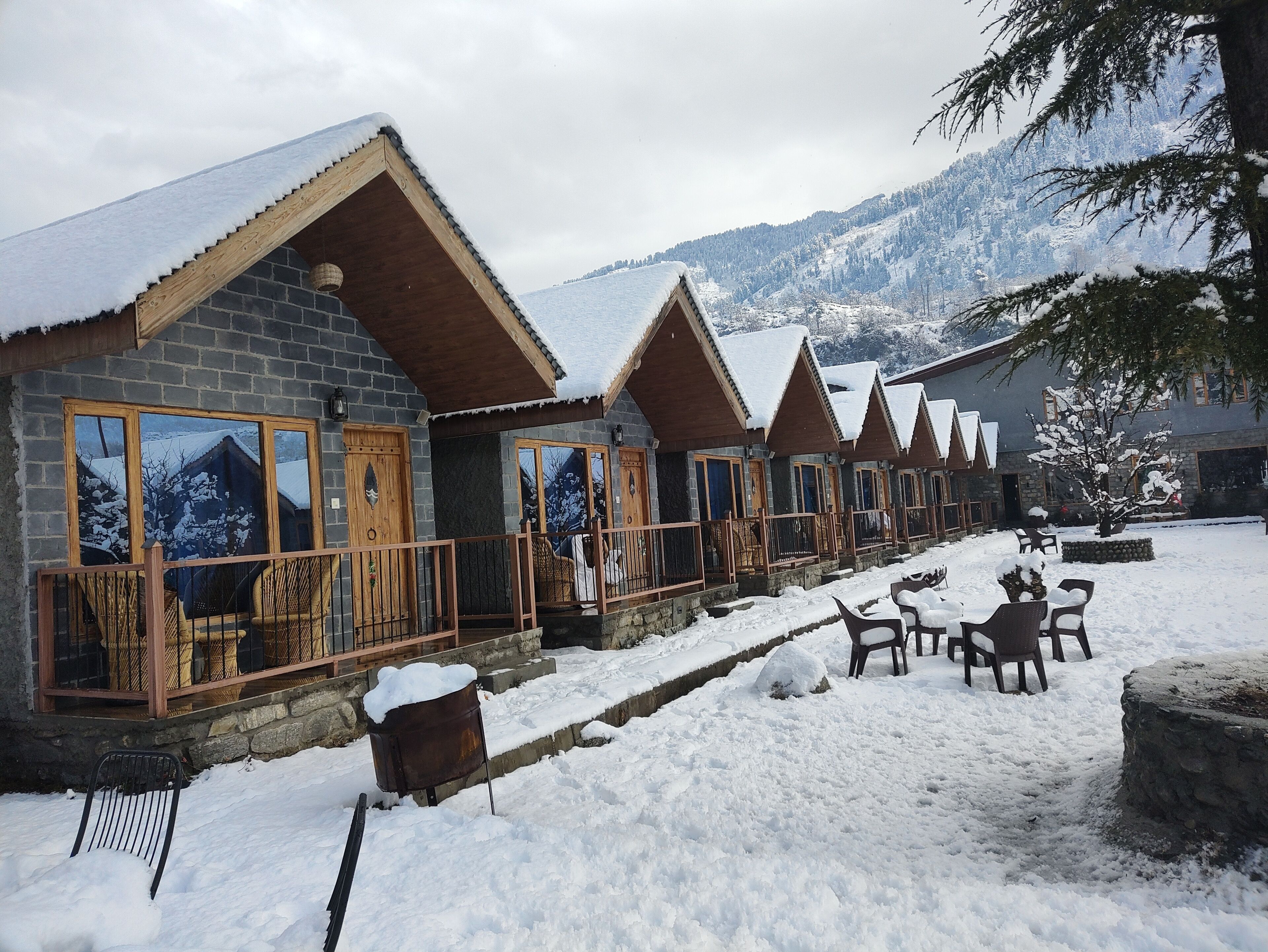 A row of wooden cabins with stone walls, each with a balcony and a view of snow-covered mountains.